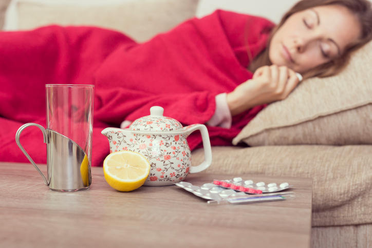 Donna influenzata sdraiata sul divano che si riposa, con in primo piano t&egrave; caldo e farmaci.