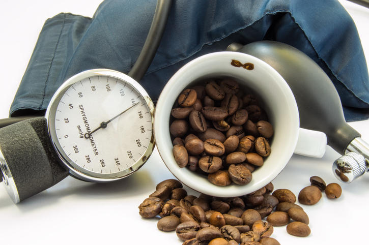 Tazza colma di chicchi di caffè, con a fianco uno strumento per la misura della pressione sanguigna (sfigmomanometro)