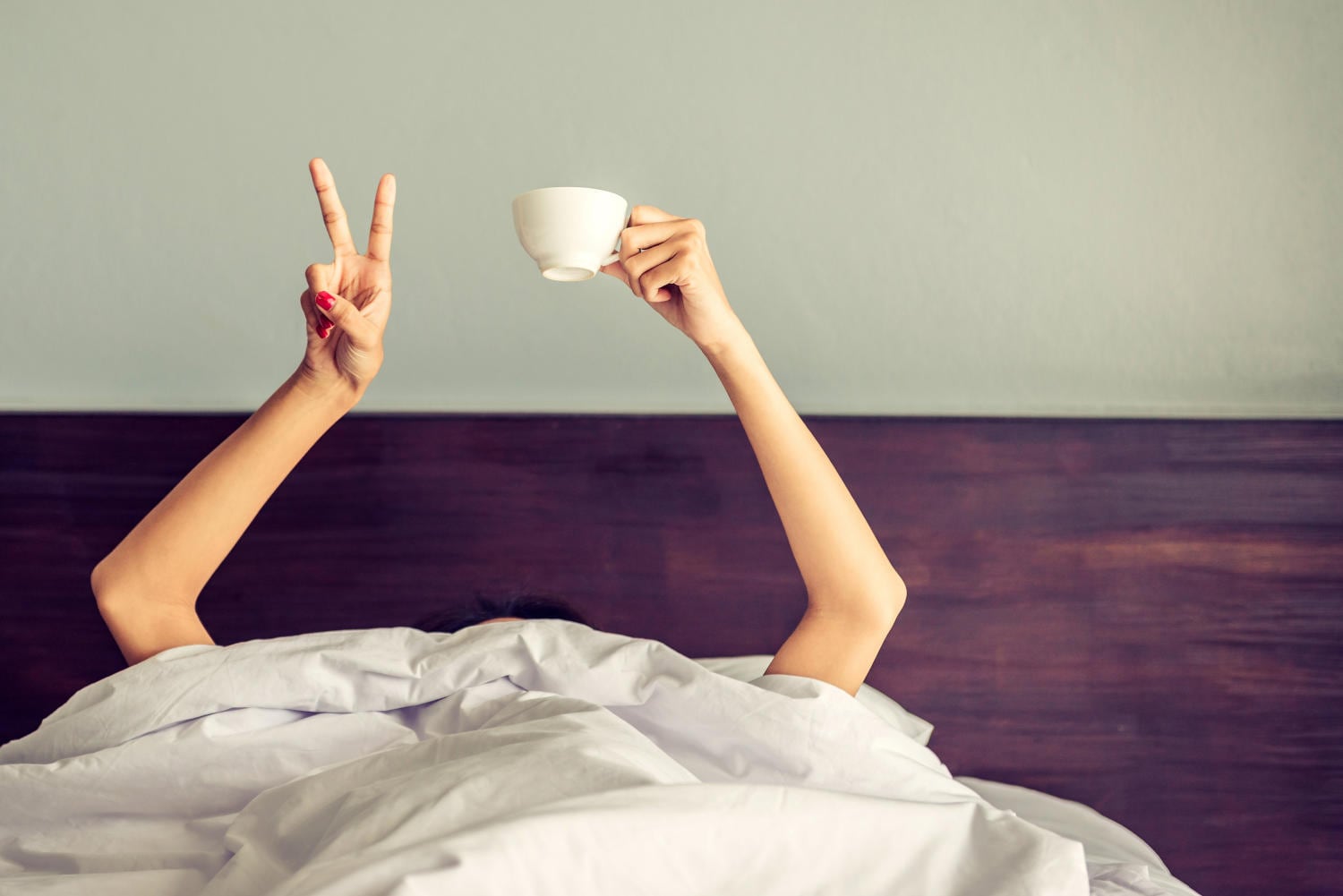 Ragazza sotto le coperte, si vedono solo braccia e mani (segno di vittoria e tazza di caffè)