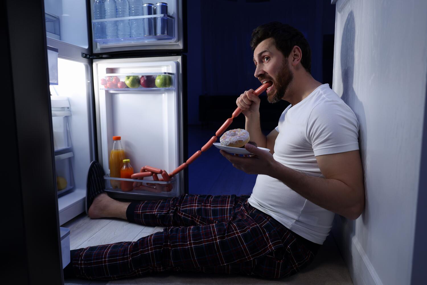 Uomo che mangia seduto davanti al frigo mordendo una salsiccia